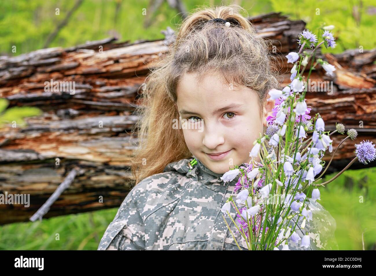 Cute Caucasian girl in camouflage and with a bouquet of forest flowers. Hiking in the forest Stock Photo