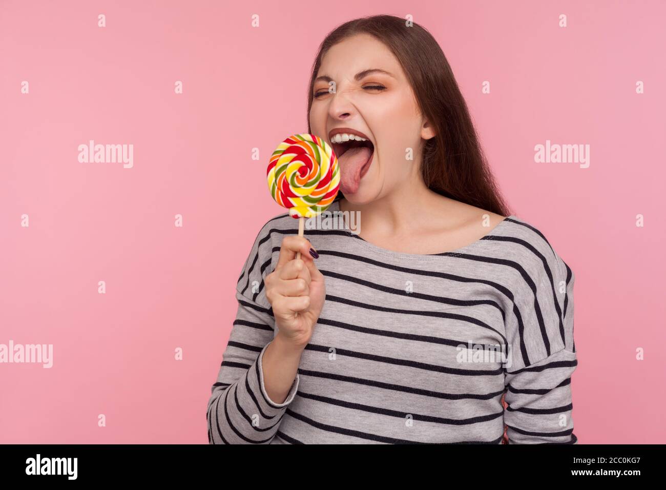 Portrait of woman in striped sweatshirt licking lollipop, tasting sweet round rainbow candy with crazy expression, enjoying delicious flavor dessert. Stock Photo