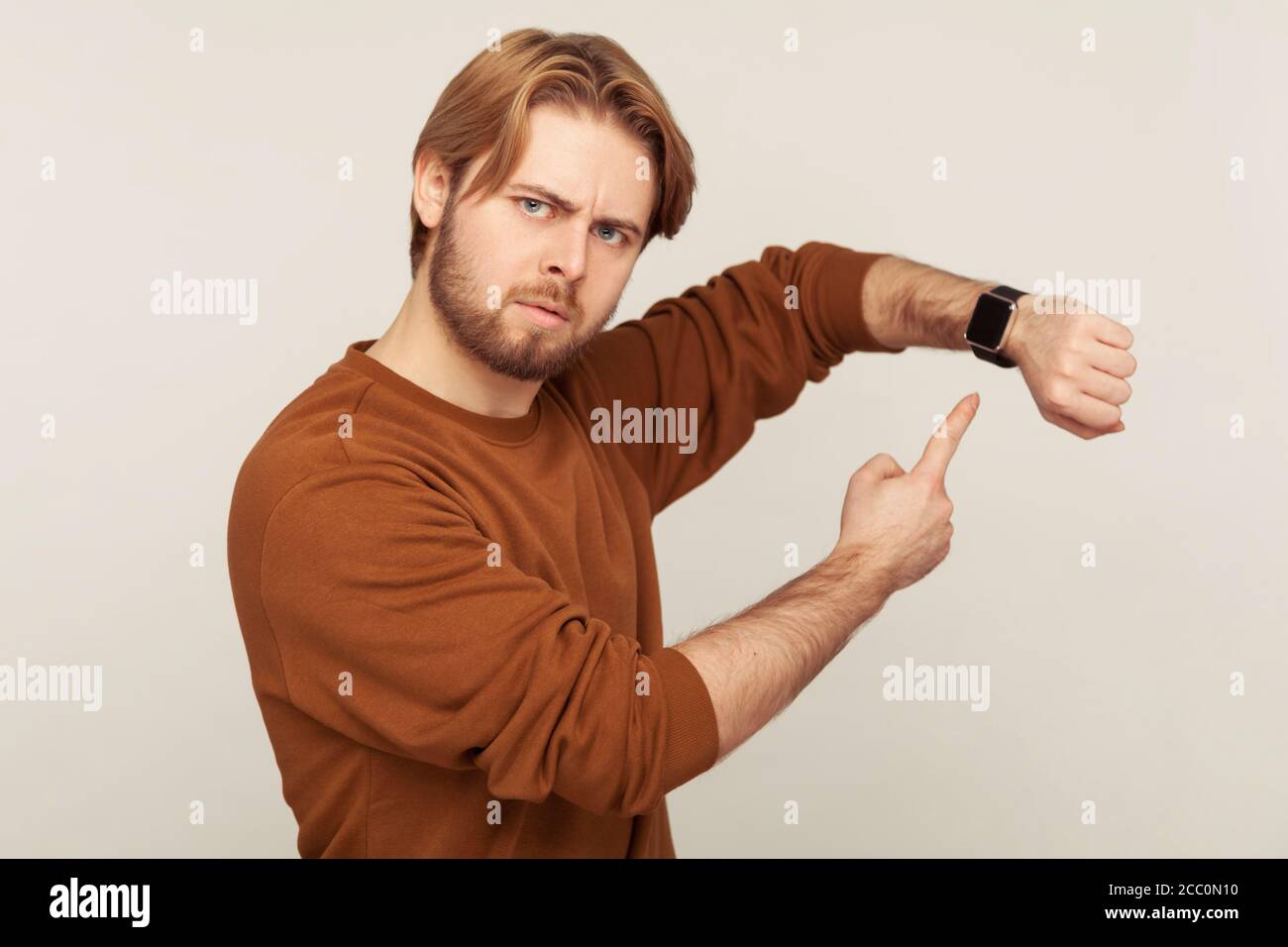 Look at time! Portrait of angry impatient man with beard in sweatshirt pointing wrist watch and looking annoyed displeased, showing clock to hurry up. Stock Photo