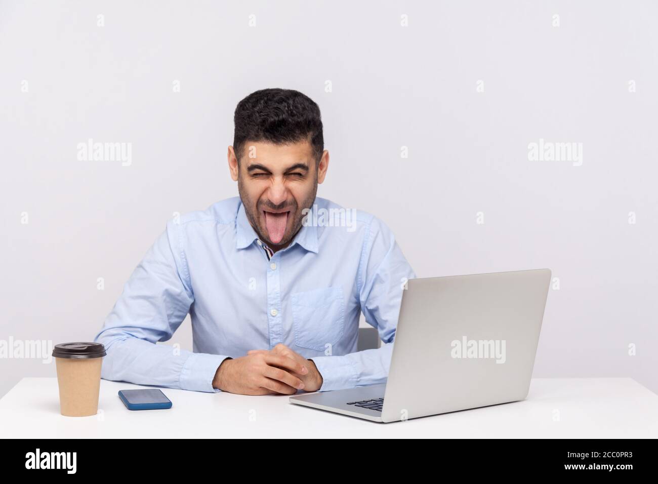 Crazy funny businessman sitting office workplace with laptop on desk, showing his tongue, looking at camera with naughty disobedient expression. indoo Stock Photo