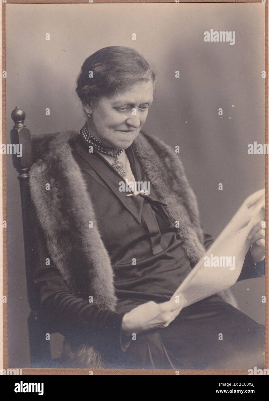 Vintage black and white photo of elegant older woman wearing a fur stole and reading newspaper 1900s Stock Photo