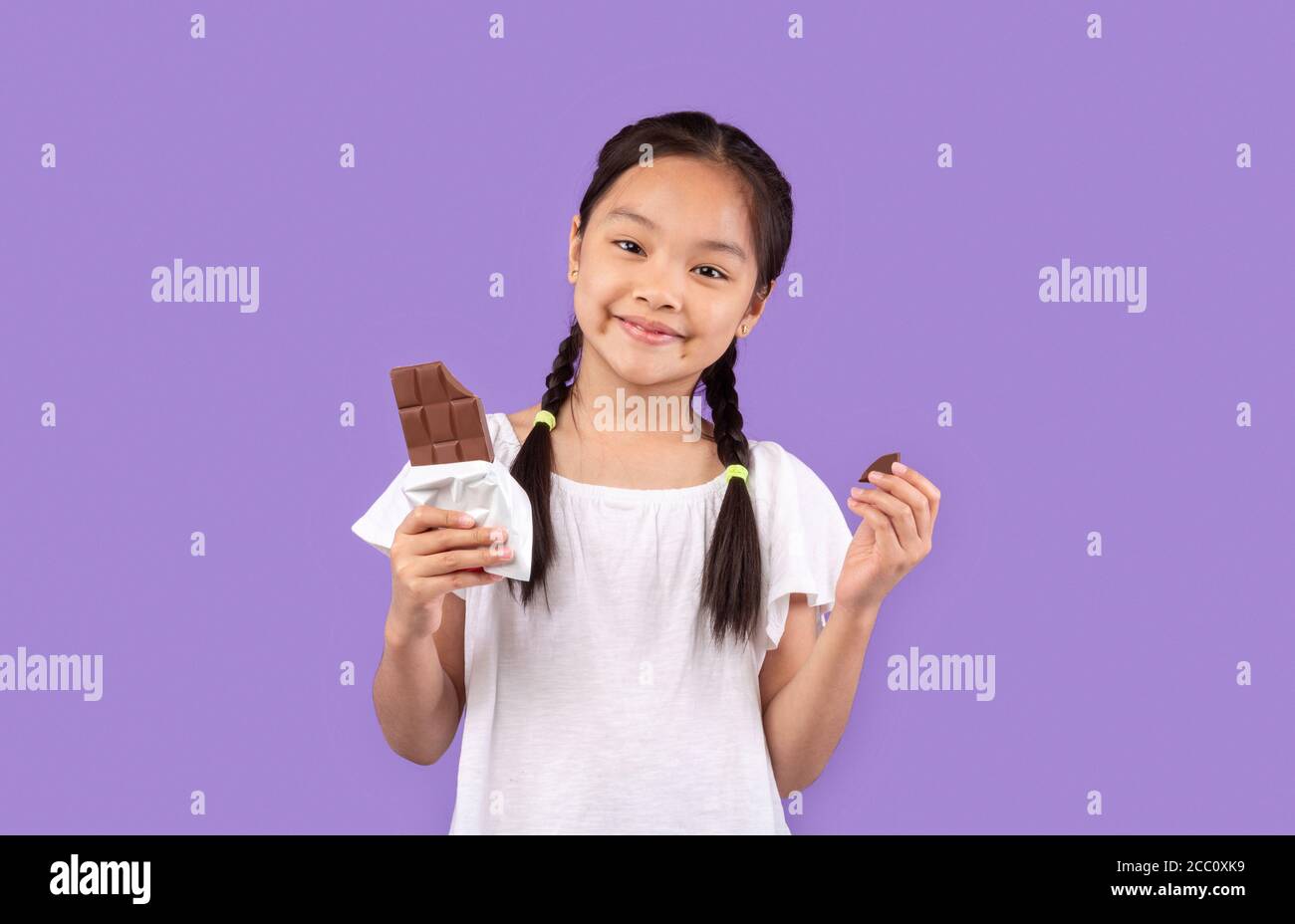 Chinese Girl Eating Chocolate Posing Over Purple Background In Studio Stock Photo