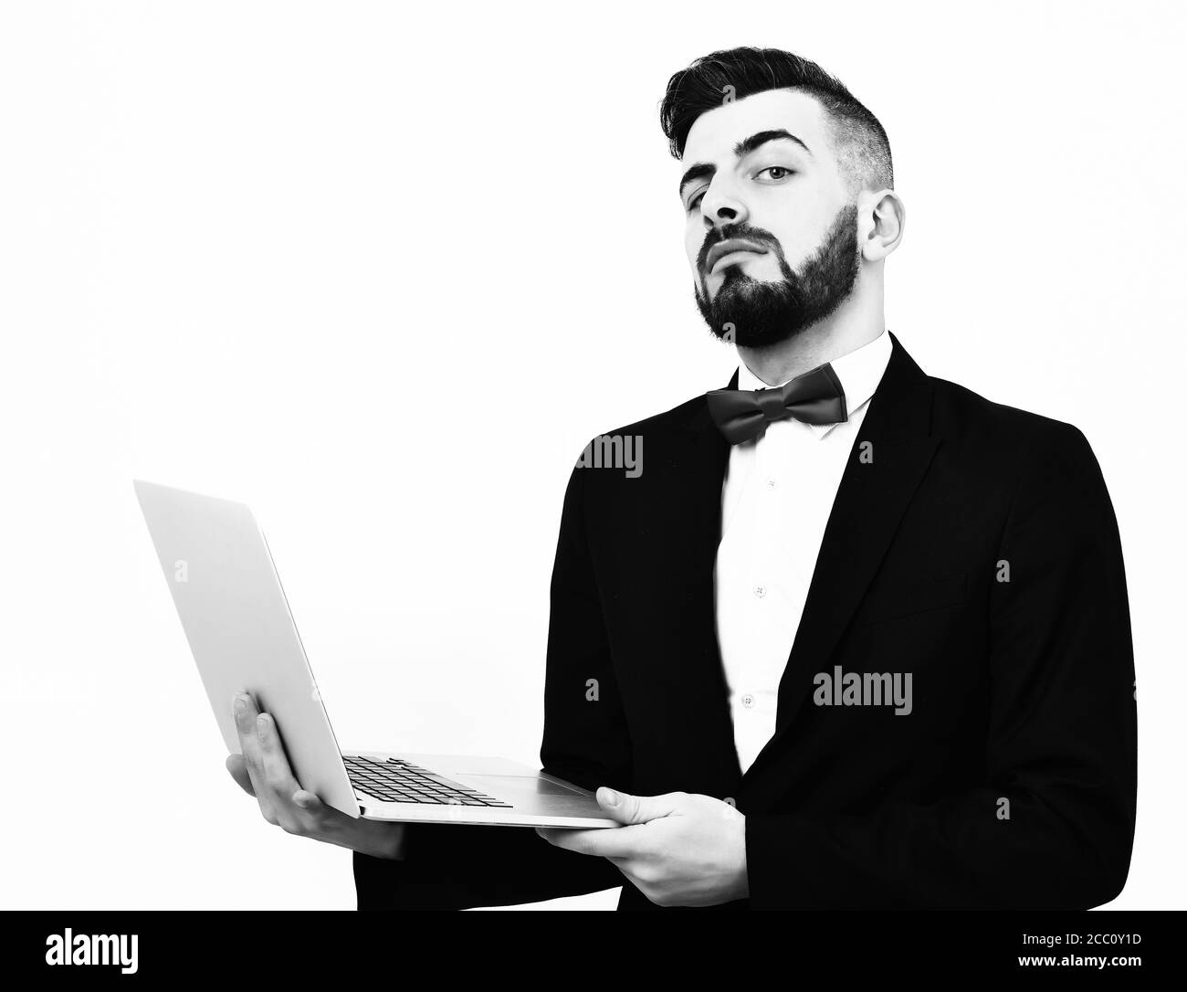 Curious and proud broker with beard in black suit and red bow holds white open laptop, isolated in white background. Concept of working online Stock Photo