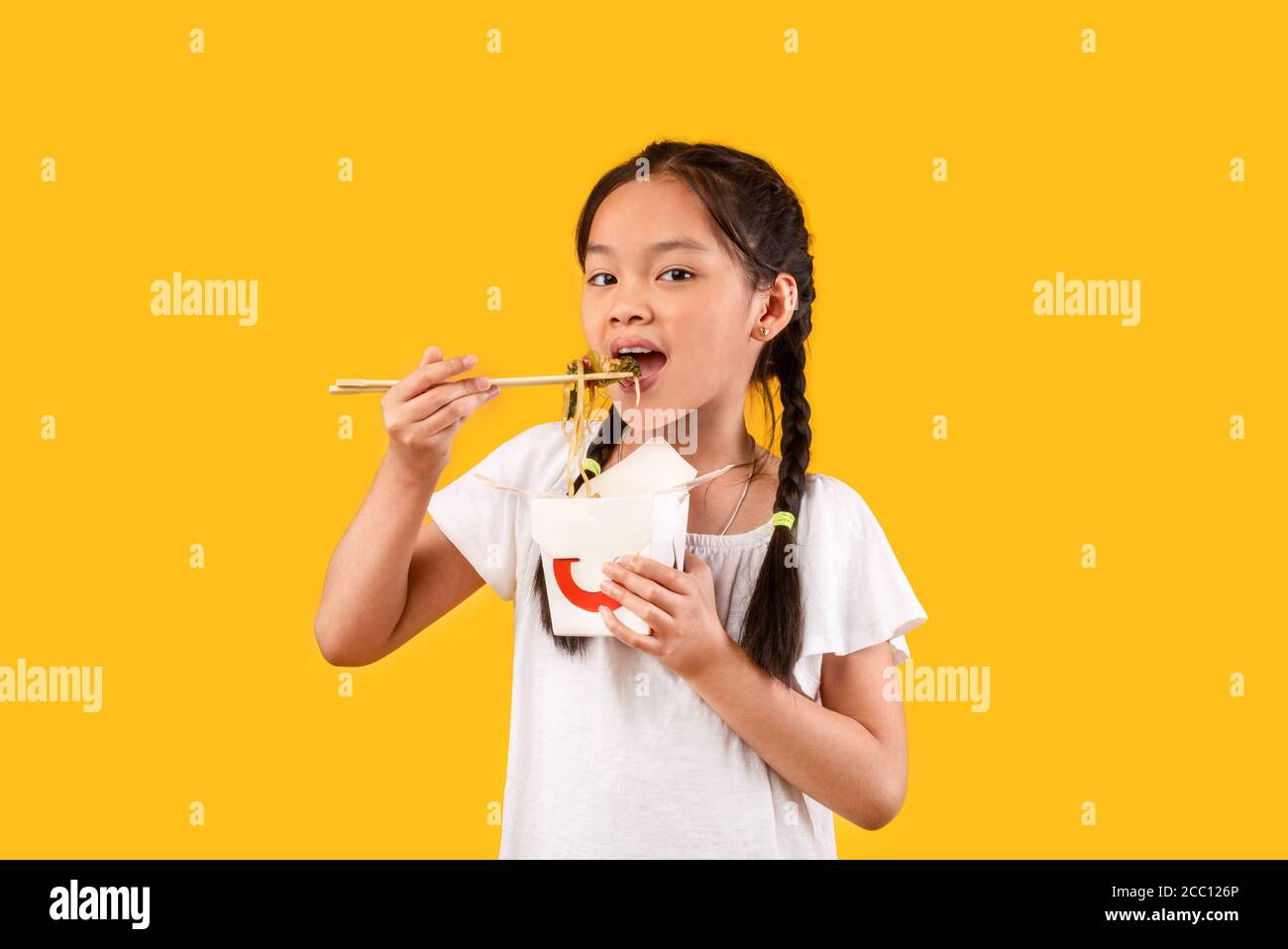 Chinese Girl Eating Noodles With Chopsticks Holding Box, Yellow Background Stock Photo