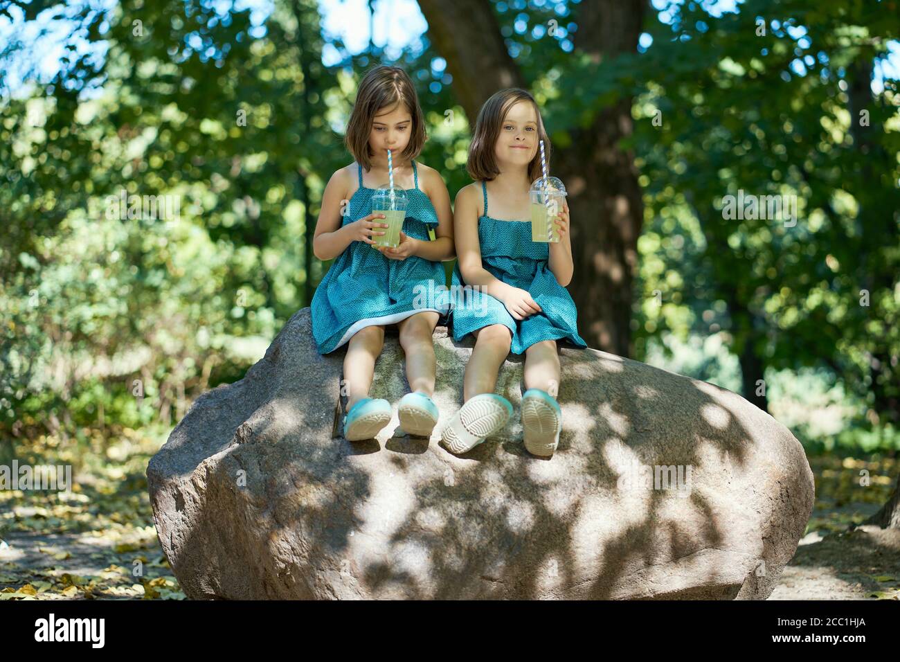 two cute little girls in dresses sit on a stone and drink lemonade  Stock Photo