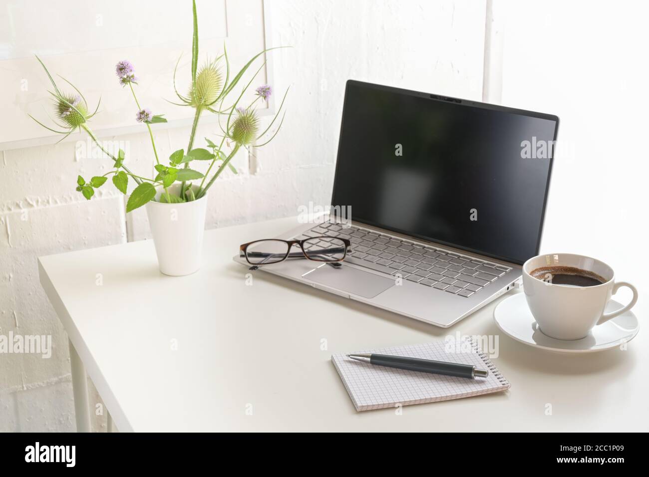 White office desk with laptop, glasses, notepad, flower bouquet and a coffee cup, small workplace at home or in a modern company against a bright wall Stock Photo