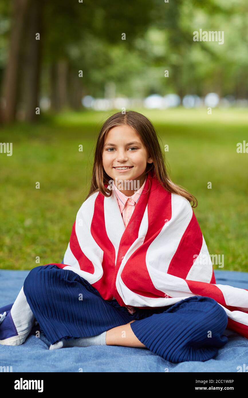 Vertical full length portrait of cute girl covered by American flag sitting on picnic blanket in park and smiling at camera, copy space Stock Photo
