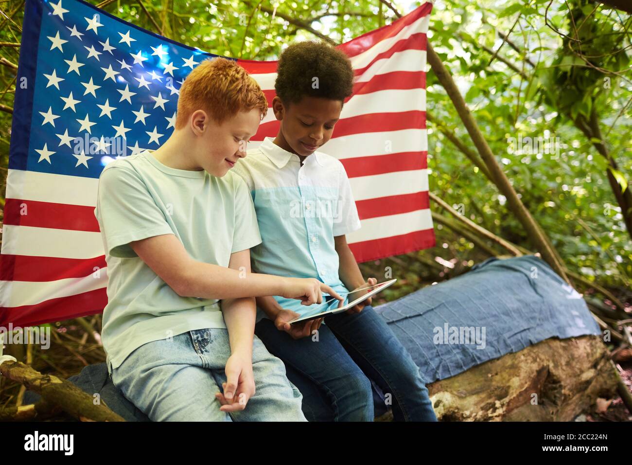 Portrait of two teenage boys using digital tablet outdoors while hiding under branches of big tree in forest or playing in back yard Stock Photo