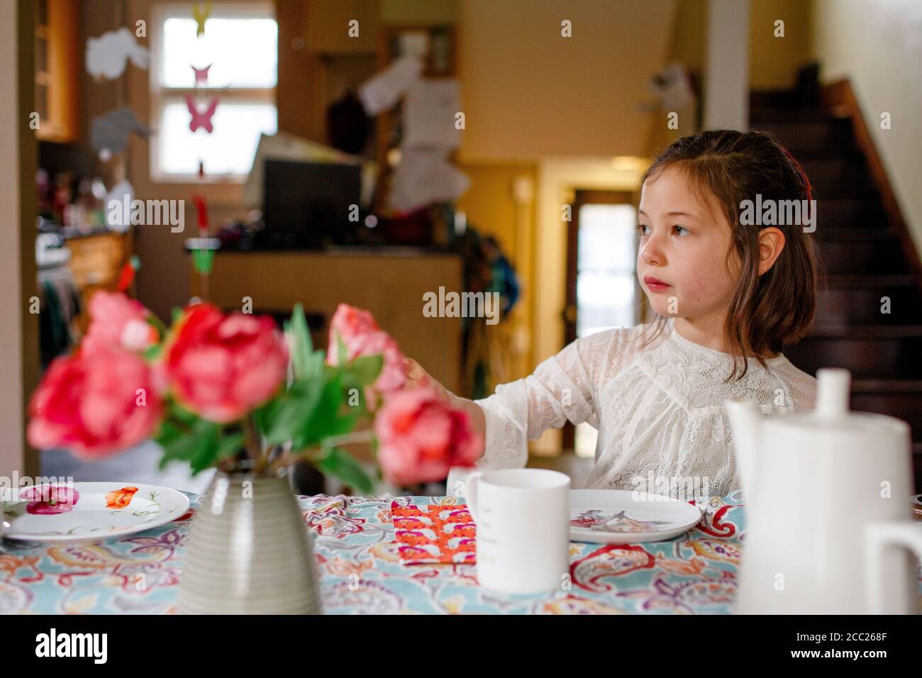 A small child in a lace dress sits alone at a table set for tea party Stock Photo