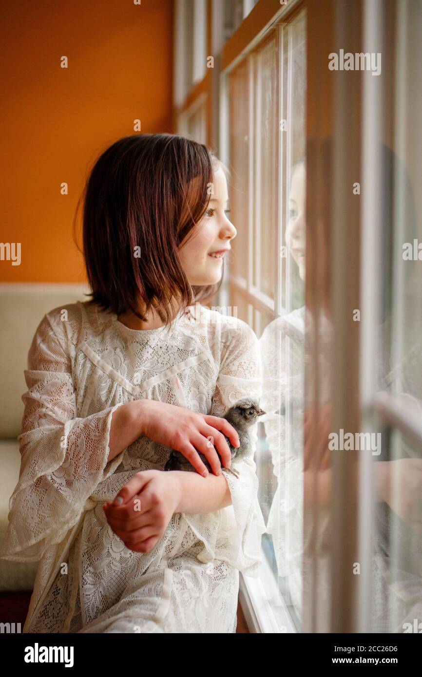 A happy girl gazes out the window, a baby chick nestled in her arms Stock Photo