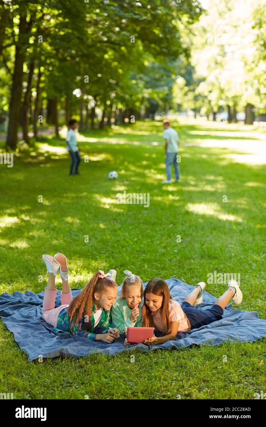 Vertical full length portrait of three teenage girls using digital tablet while lying on green grass in park outdoors lit by sunlight, copy space Stock Photo