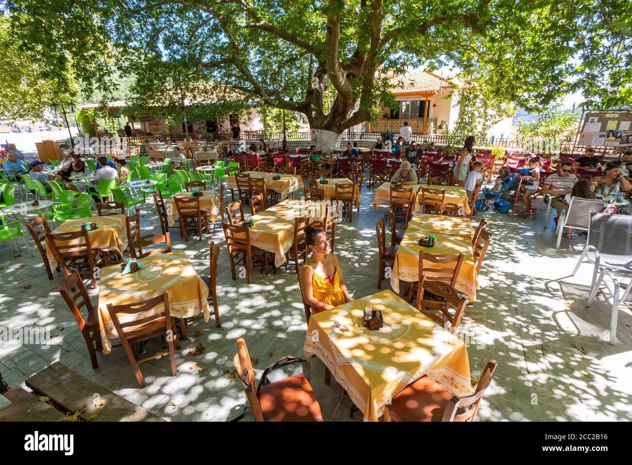 A girl sitting at a taverna table under dappled light in a square in Karya, Lefkada, Ionian Islands, Greece Stock Photo