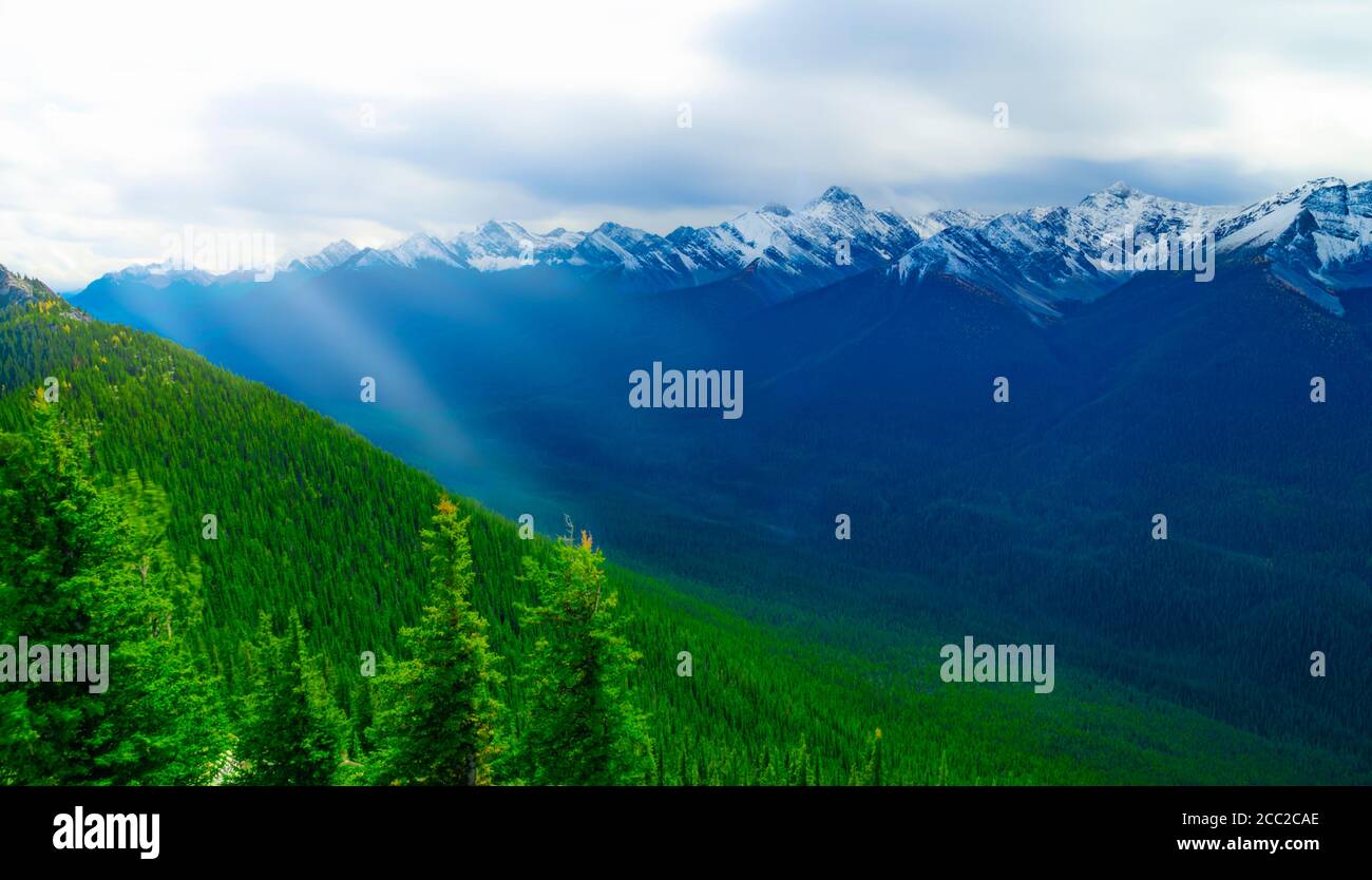 Massive Mountain Range With Snow Capped Peaks And Sun Rays Over Rich Green Forest Stock Photo