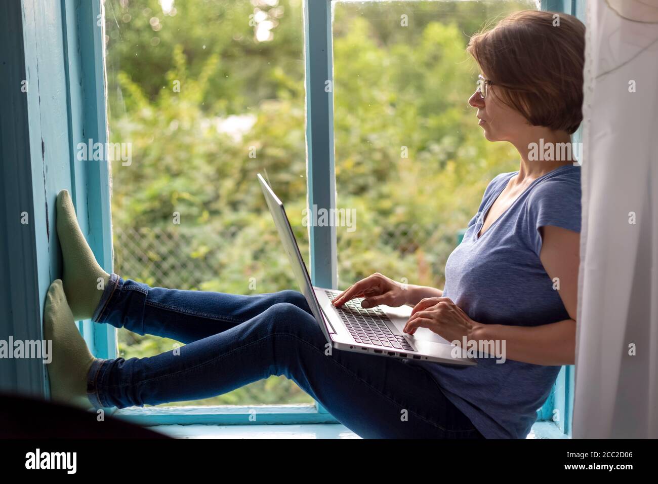 Woman freelancer sitting on a windowsill working on a laptop Stock Photo