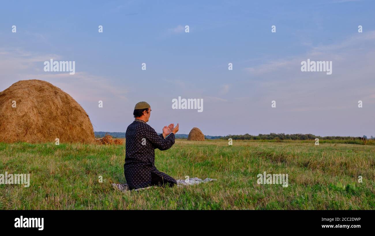 A Muslim senior man wearing a skullcap and traditional clothes prays in an hayfield. Stock Photo