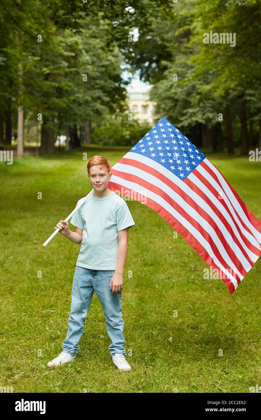Vertical full length portrait of smiling red haired boy carrying American flag while standing outdoors in park, copy space Stock Photo