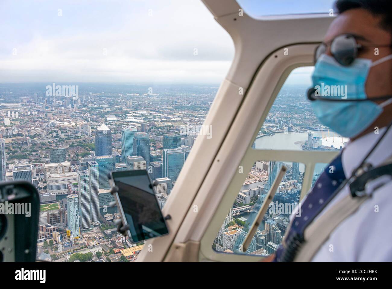Horizontal portrait of a pilot wearing a face mask flying a helicopter over Canary Wharf in London. Stock Photo