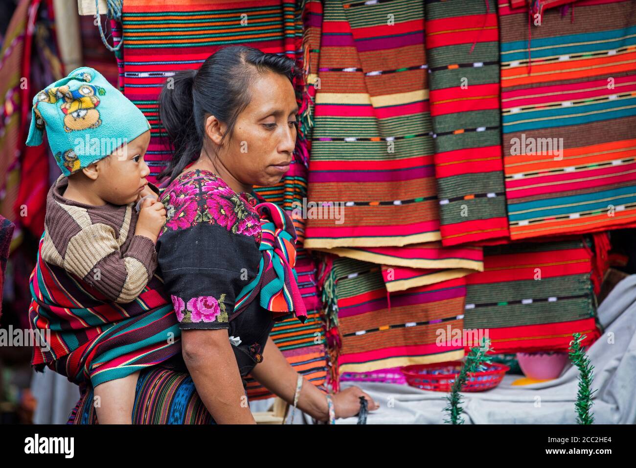 Local Mayan K'iche woman with child on her back selling fabric on market day in the town Chichicastenango, El Quiché, Guatemala, Central America Stock Photo