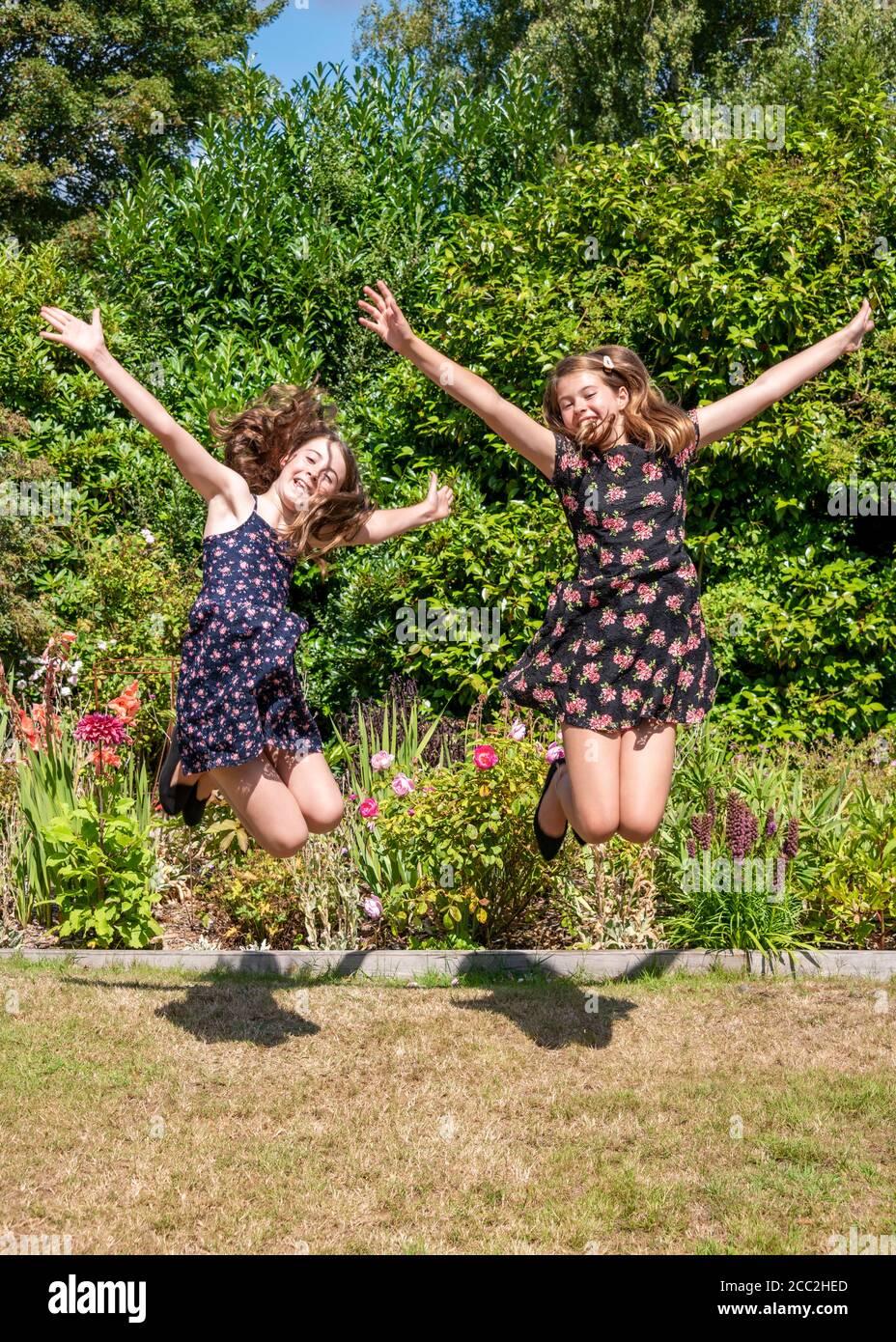 Vertical portrait of two young girls jumping with excitement with their arms outstretched in the garden. Stock Photo