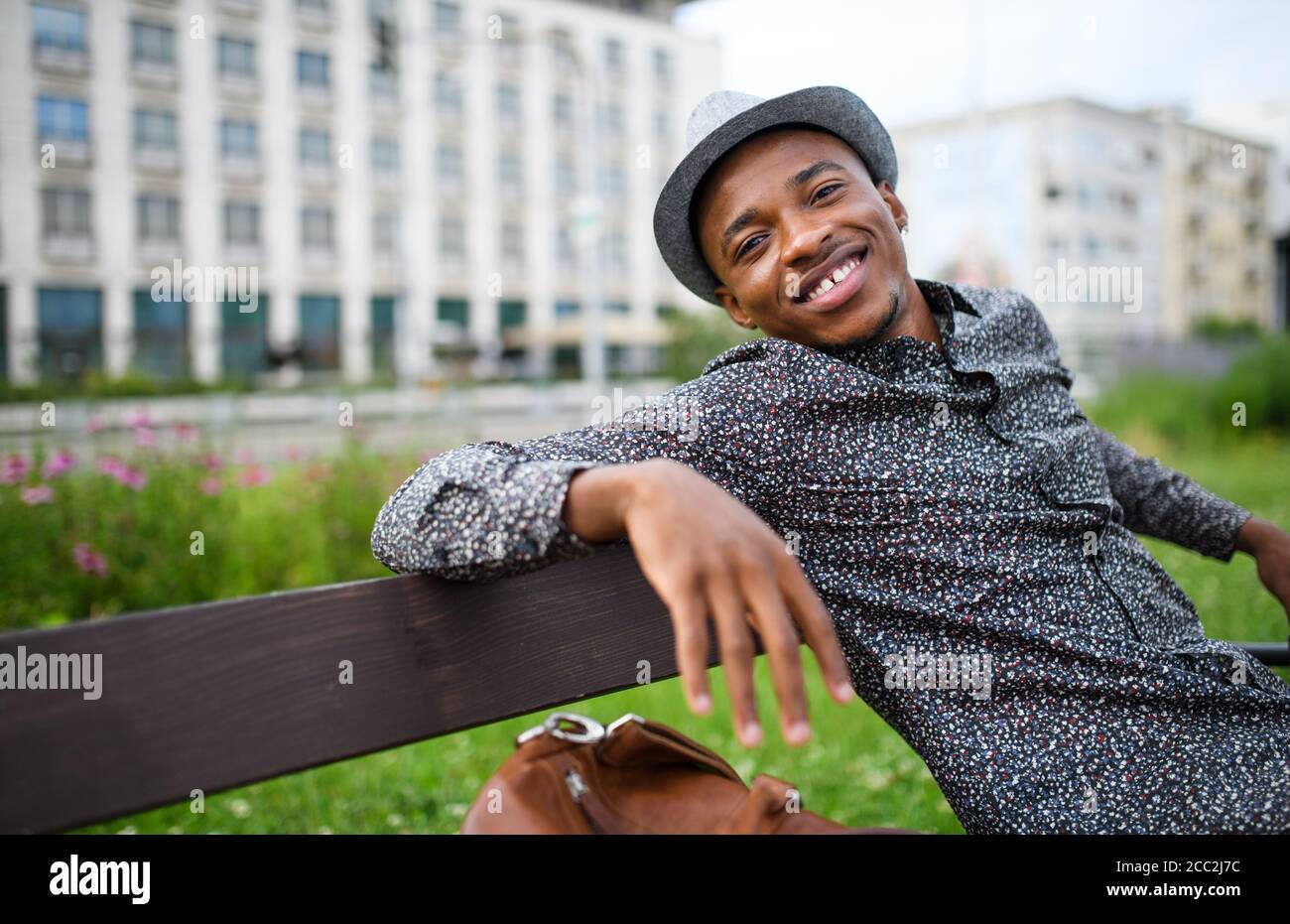 Cheerful young black man commuter sitting on bench outdoors in city. Stock Photo