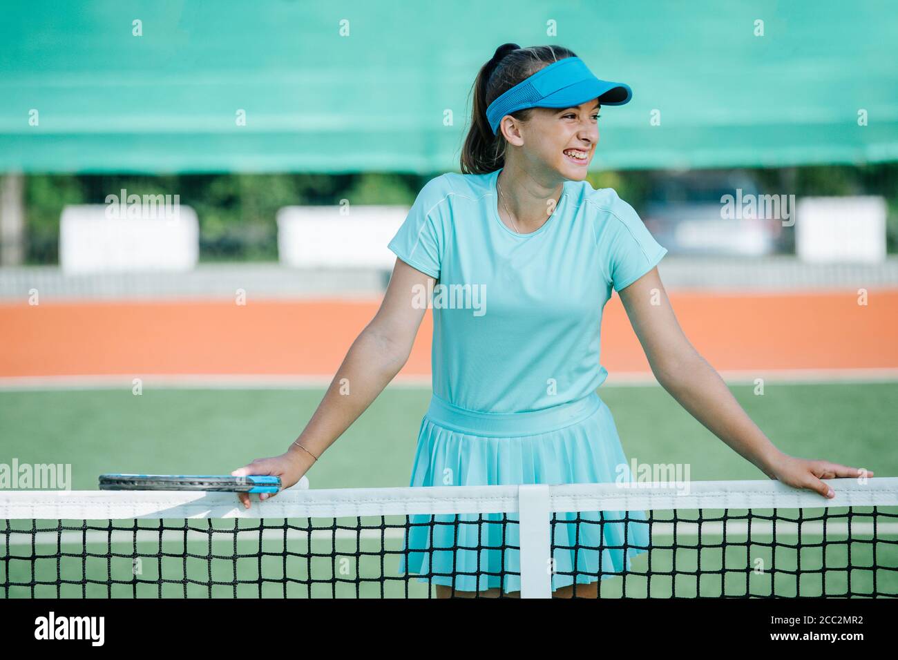 Grinning teenage girl standing on tennis court, leaning on the net, resting Stock Photo