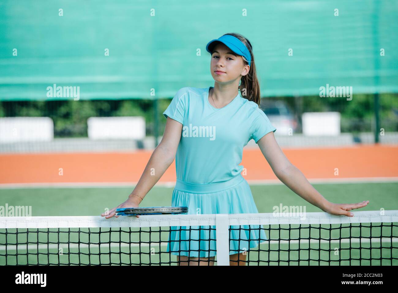 Portrait of a teenage girl standing on tennis court, leaning on the net, resting Stock Photo