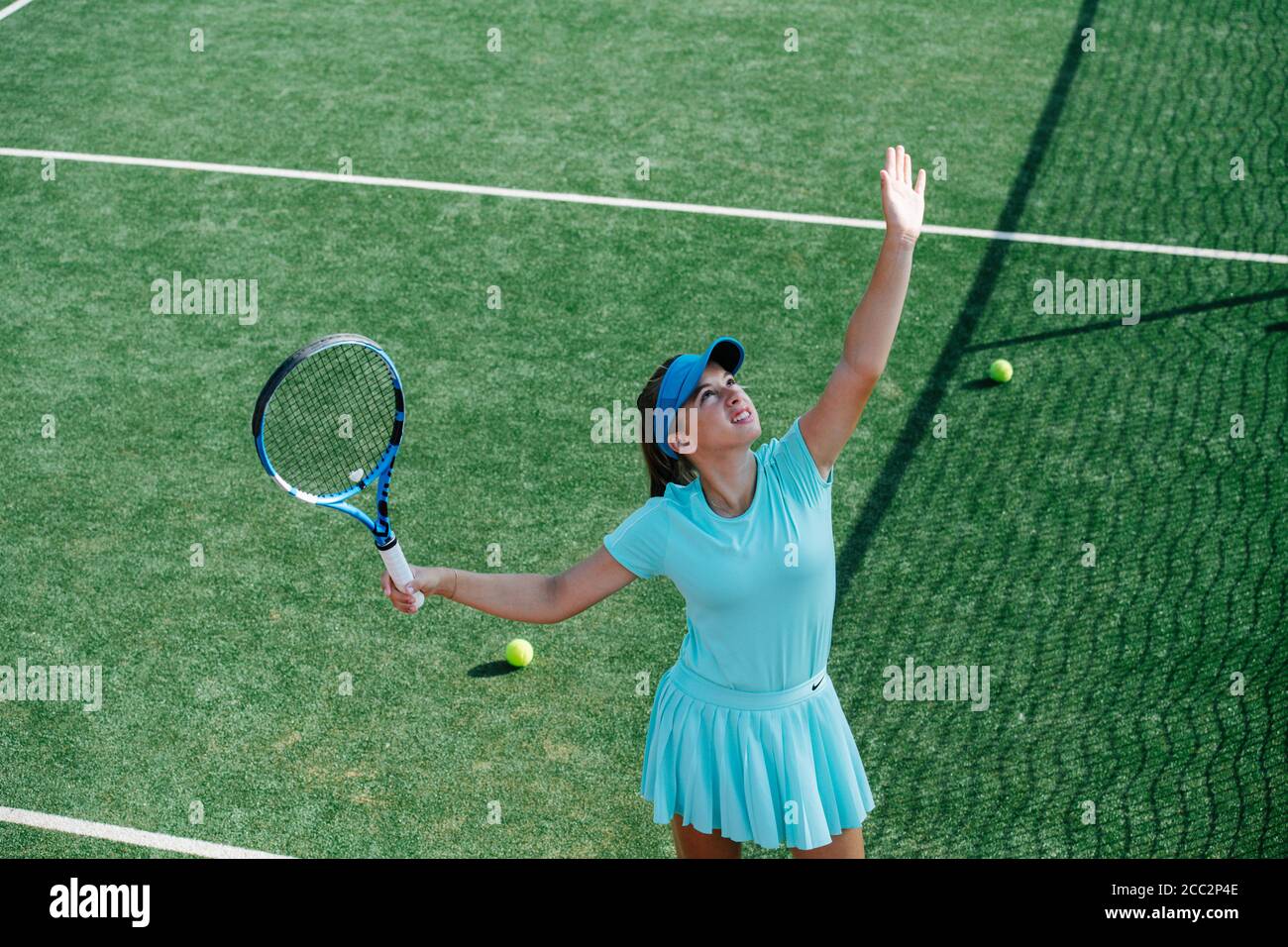 Squinting young girl training on a tennis court, throwing ball up and serving Stock Photo