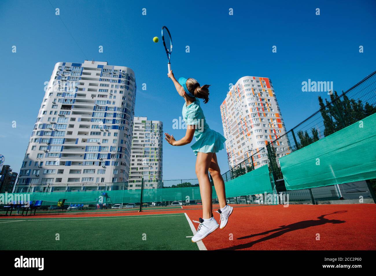 Jumping teenage girl training on a tennis court, throwing ball up and serving Stock Photo