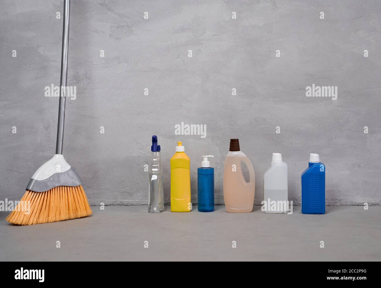 Home cleaning products and tools. Broom, bottles with different detergents standing on the floor against grey wall. Housework, house cleaning and disinfection, housekeeping Stock Photo