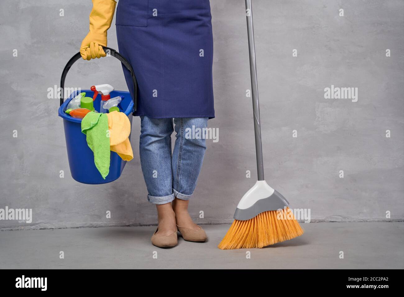 Cleaning the house. Cropped shot of woman in uniform and yellow rubber gloves holding broom and bucket different cleaning products while standing against grey wall. Housekeeping, housework, cleaning Stock Photo
