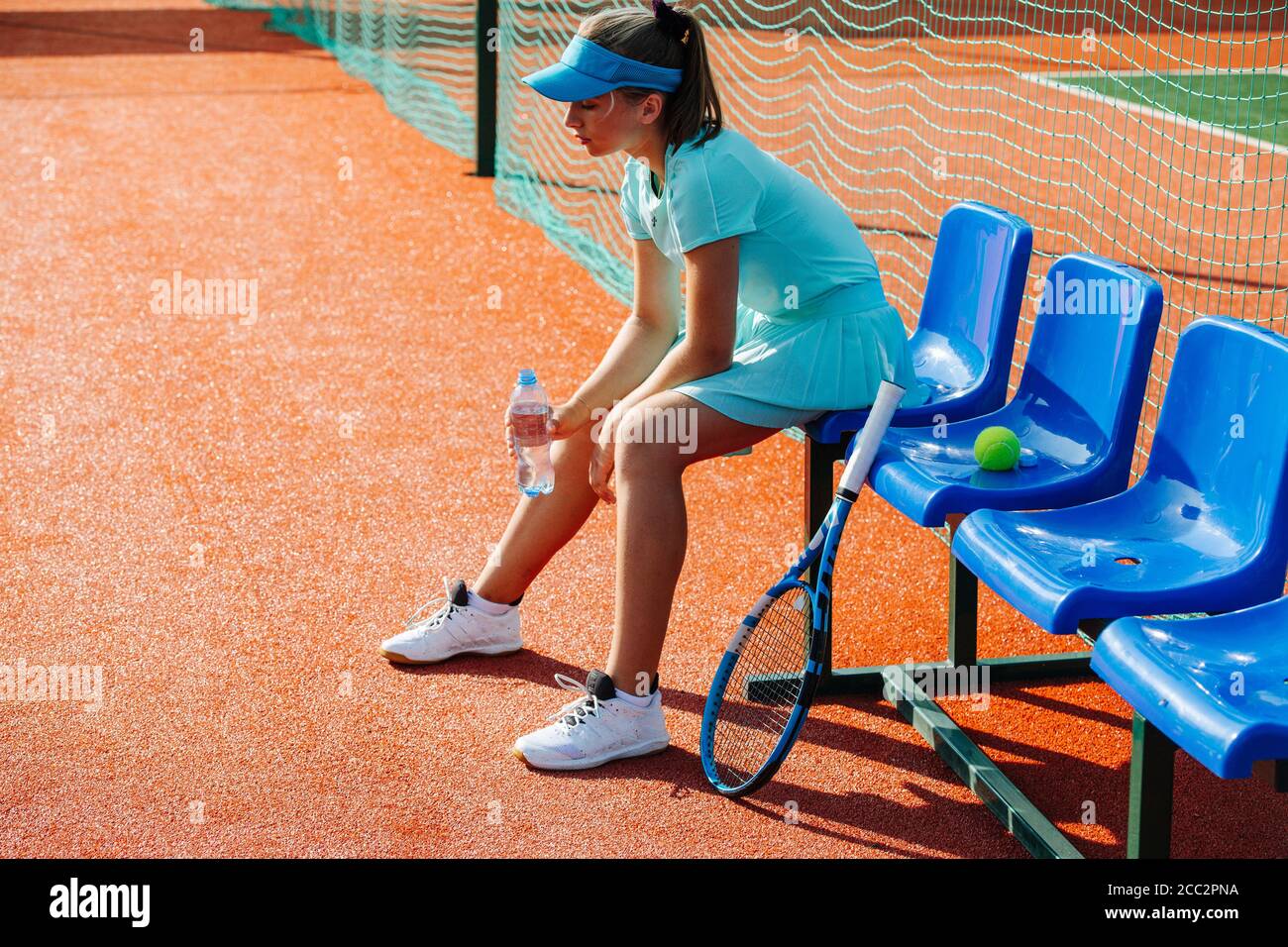 Tired, spent girl sitting on a chair bench next to tennis court, taking break Stock Photo