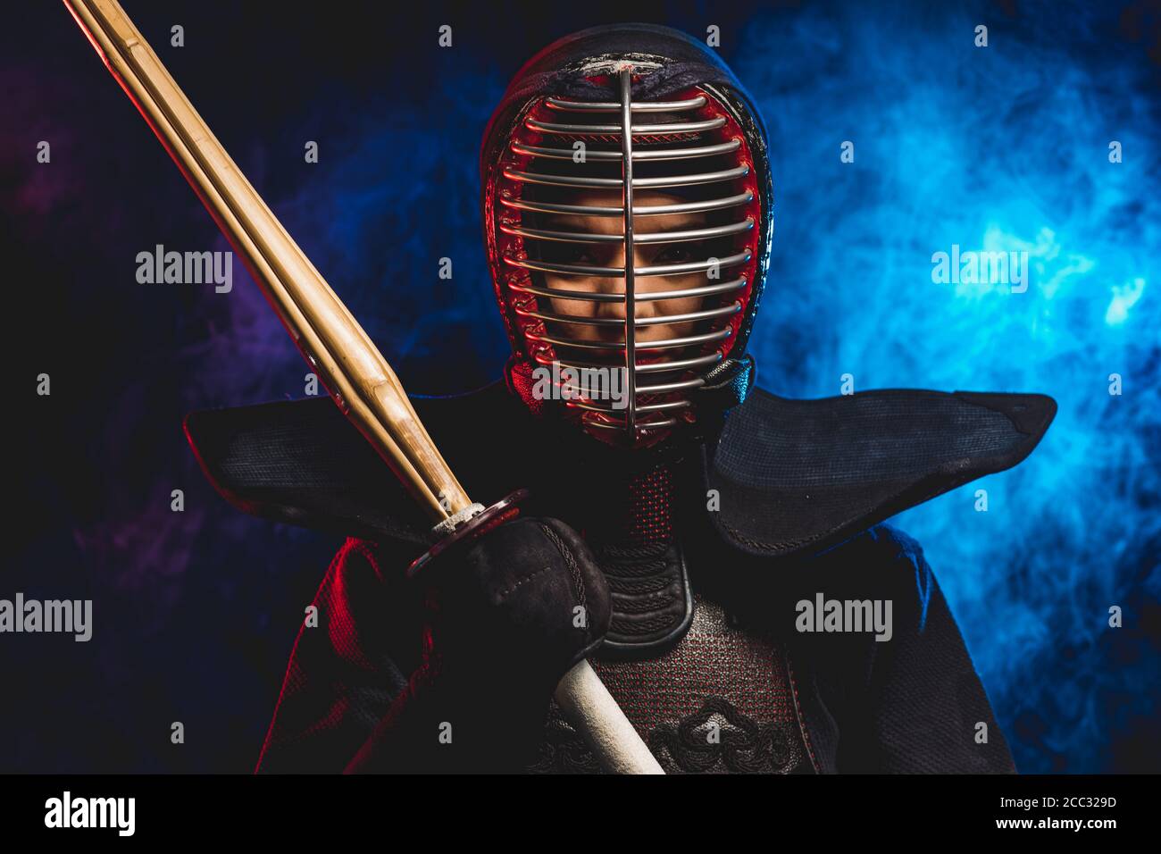 portrait of kendo fighter in uniform and protective helmet. combat warrior holding shinai in hand bamboo sword isolated over smoky space Stock Photo