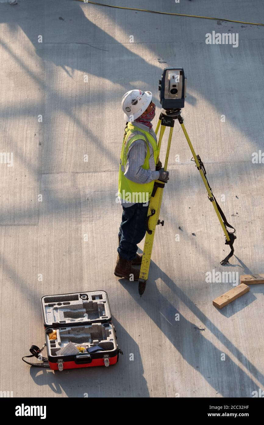 Construction worker wearing safety gear and face covering uses tripod-mounted transit on parking garage job site near downtown Austin, Texas. Stock Photo