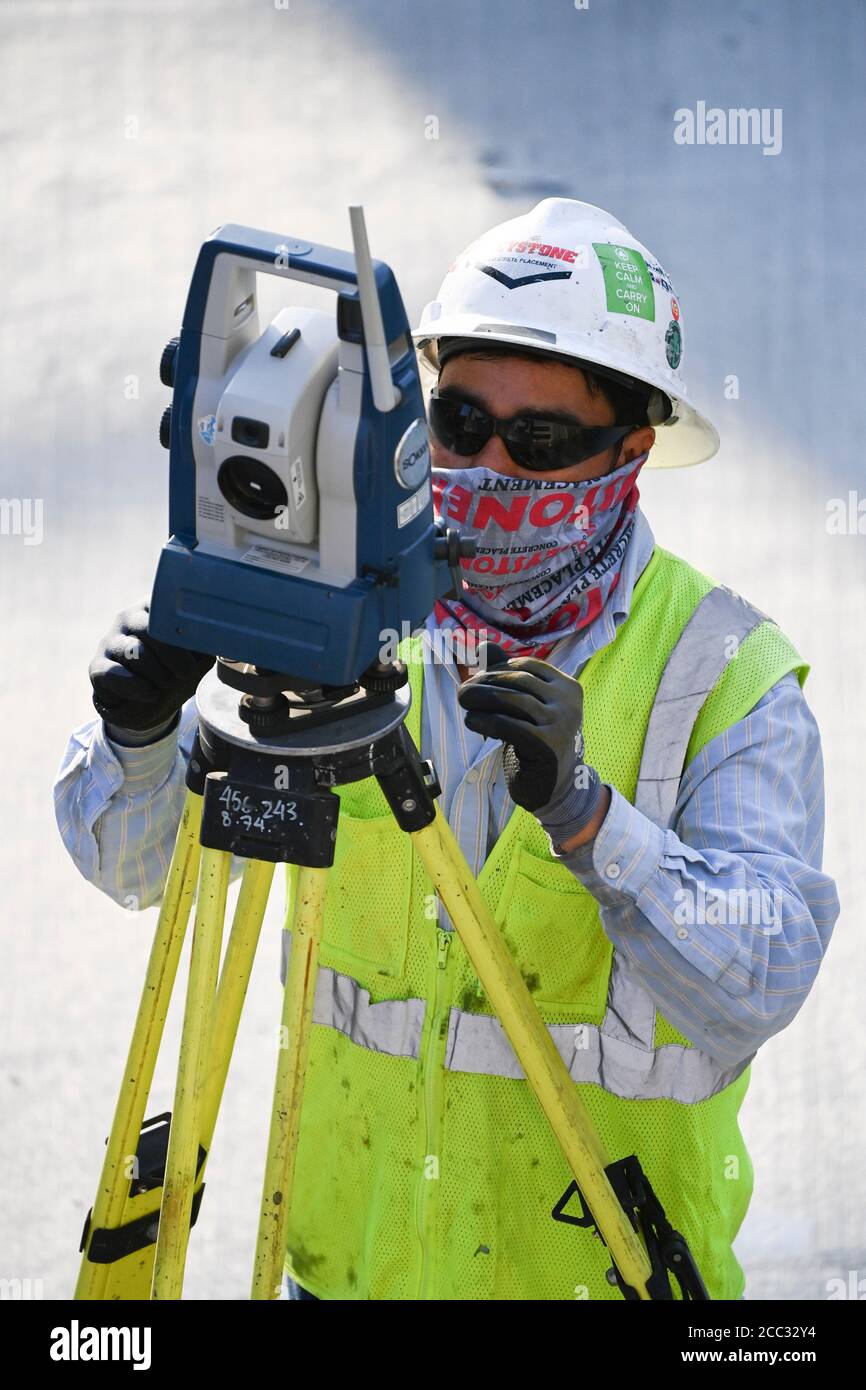 Construction worker wearing safety gear and face covering uses tripod
