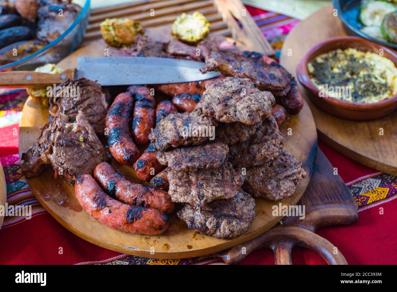 A mixture of grilled meats on a wood board. Barbecue meat on a table with a Peruvian style table cloth. Stock Photo