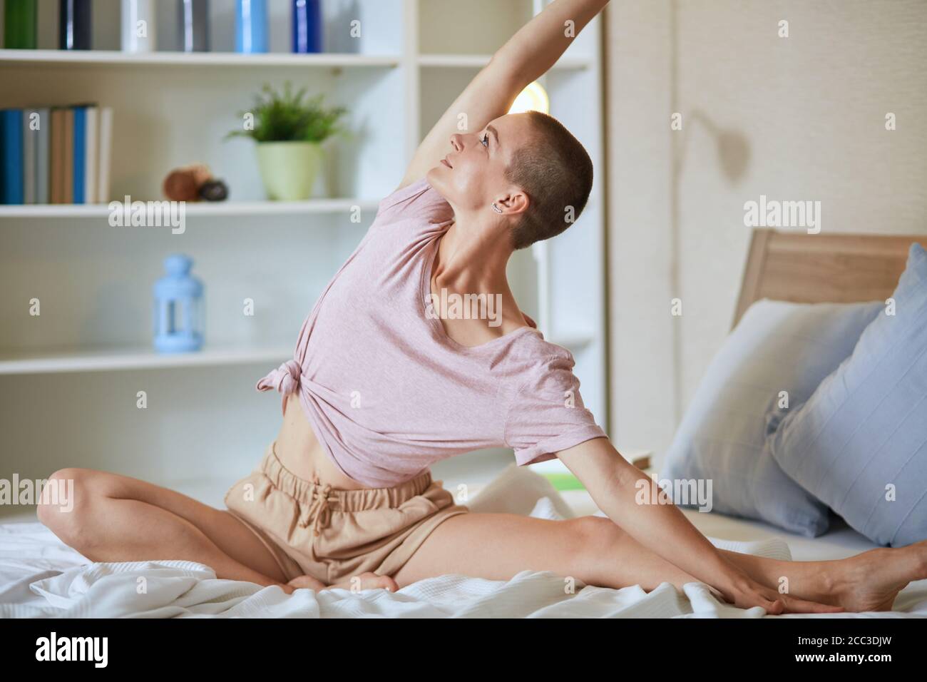 Caucasian woman on bed practice yoga exercises, background white room. Hands raised up. Stock Photo