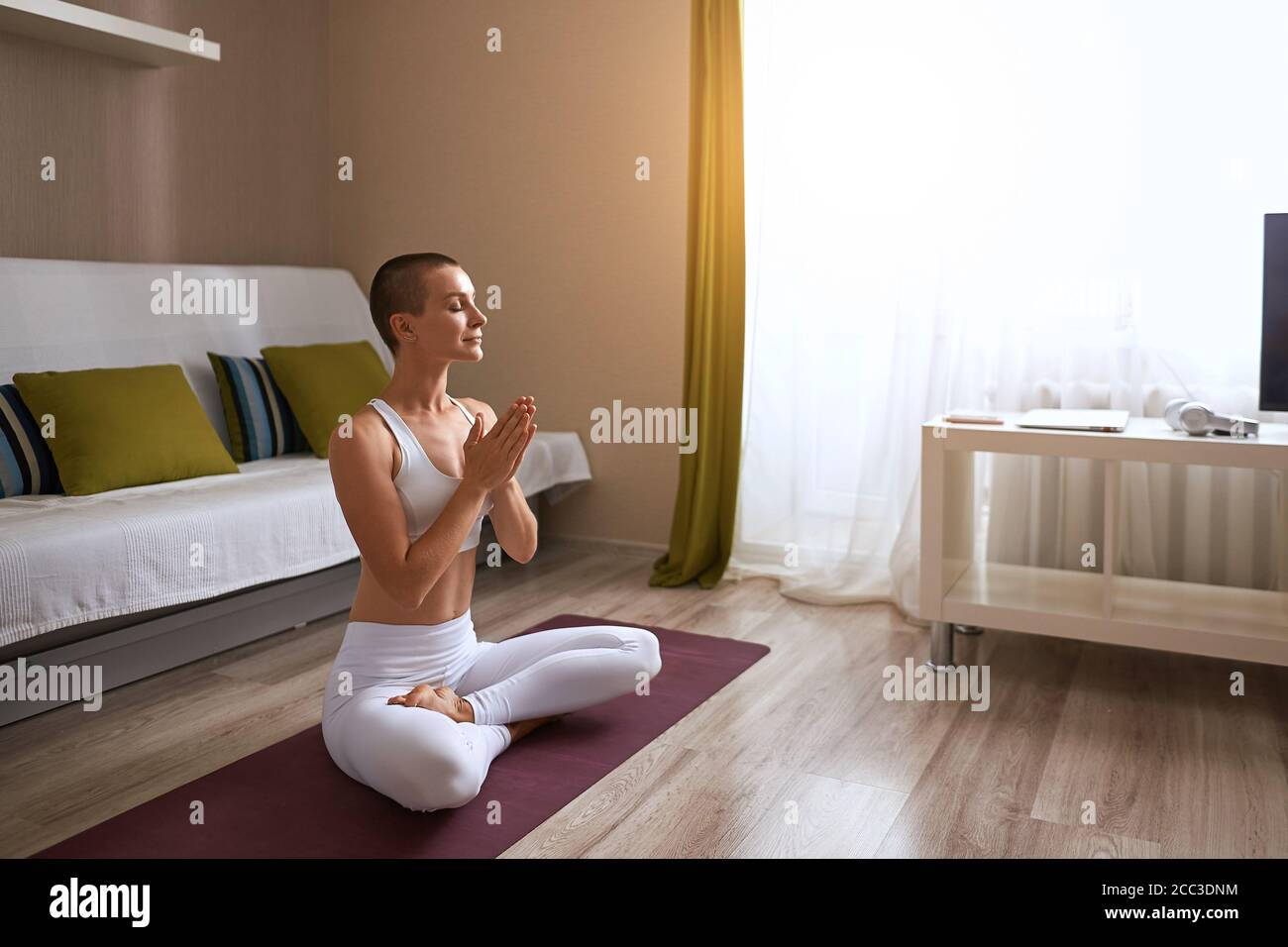 Quiet caucasian woman with closed eyes keeping calm at home. Pray. Doing exercise sitting on purple mat. Background window, bright room Stock Photo