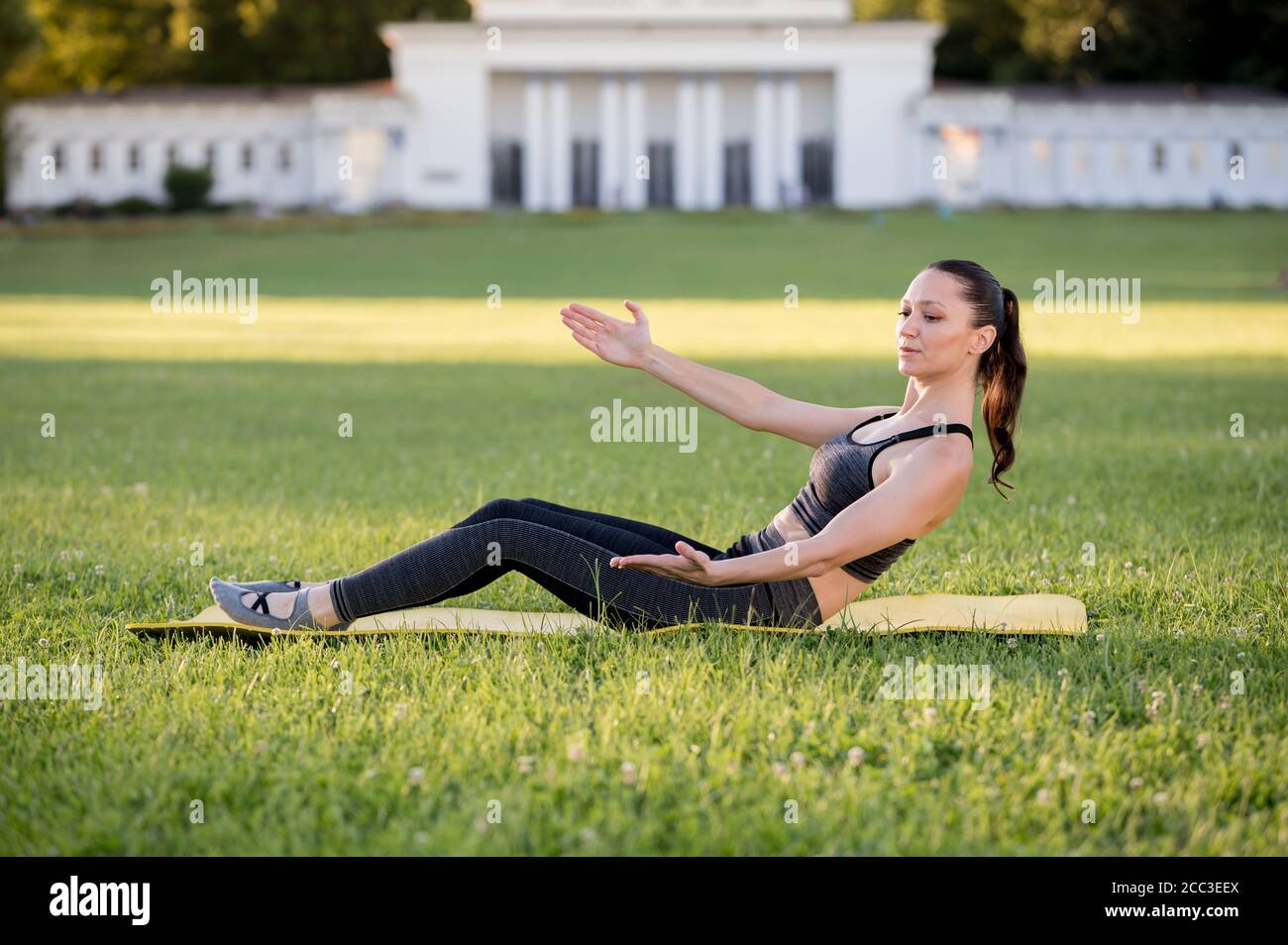 Beautiful young woman lying on a yellow mattress, pose while wearing a tight sports outfit in the park doing pilates or yoga, roll up intermediate exe Stock Photo