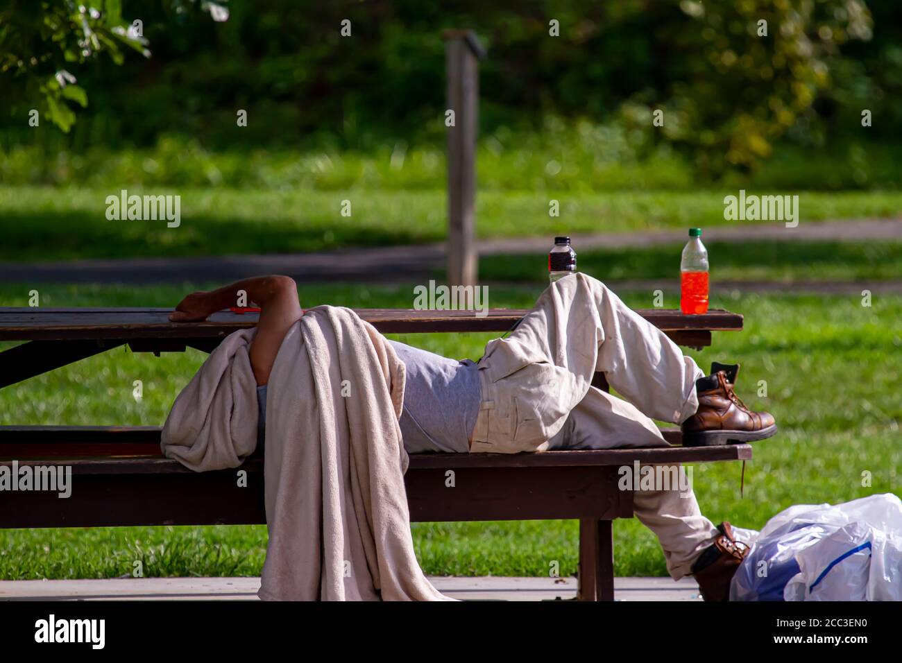 A homeless caucasian man is sleeping on a picnic table in a shaded area of a park. He is covering his head with a dirty blanket. His belongings are in Stock Photo