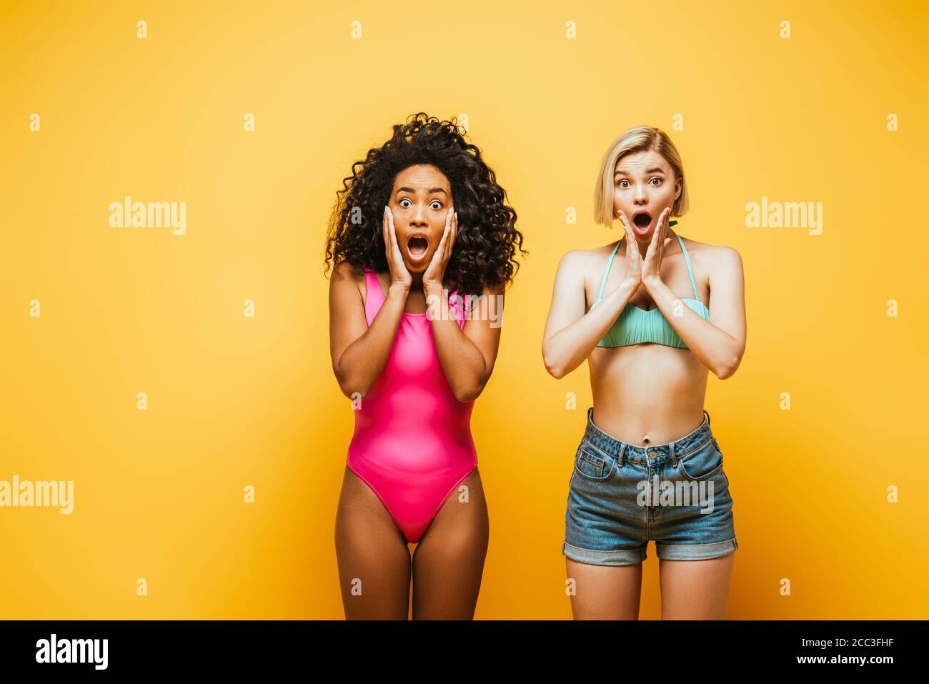 shocked multicultural women in summer outfit touching faces on yellow Stock Photo