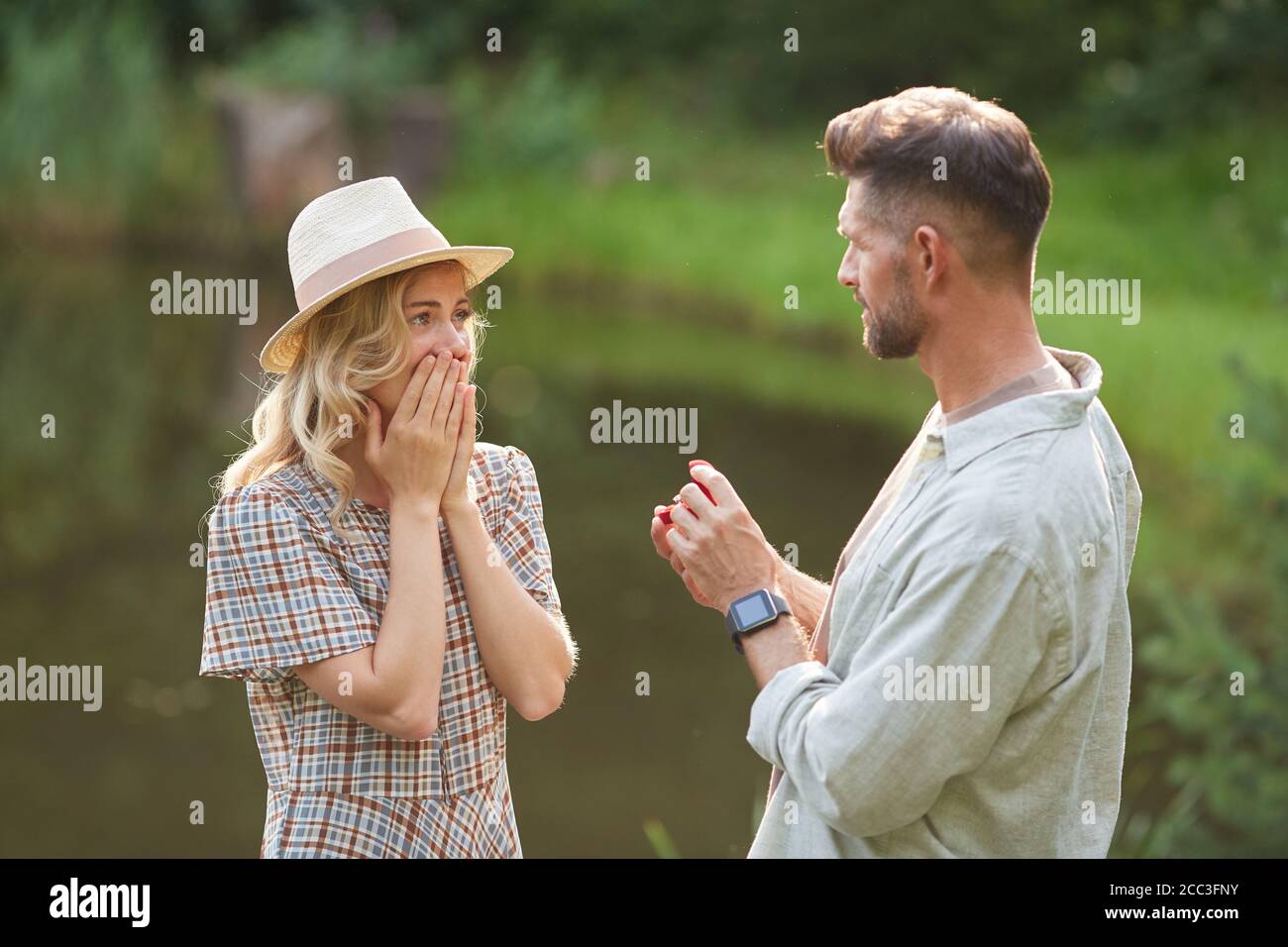 Waist up portrait of man proposing to beautiful girlfriend while standing by lake outdoors in sunlight, copy space Stock Photo