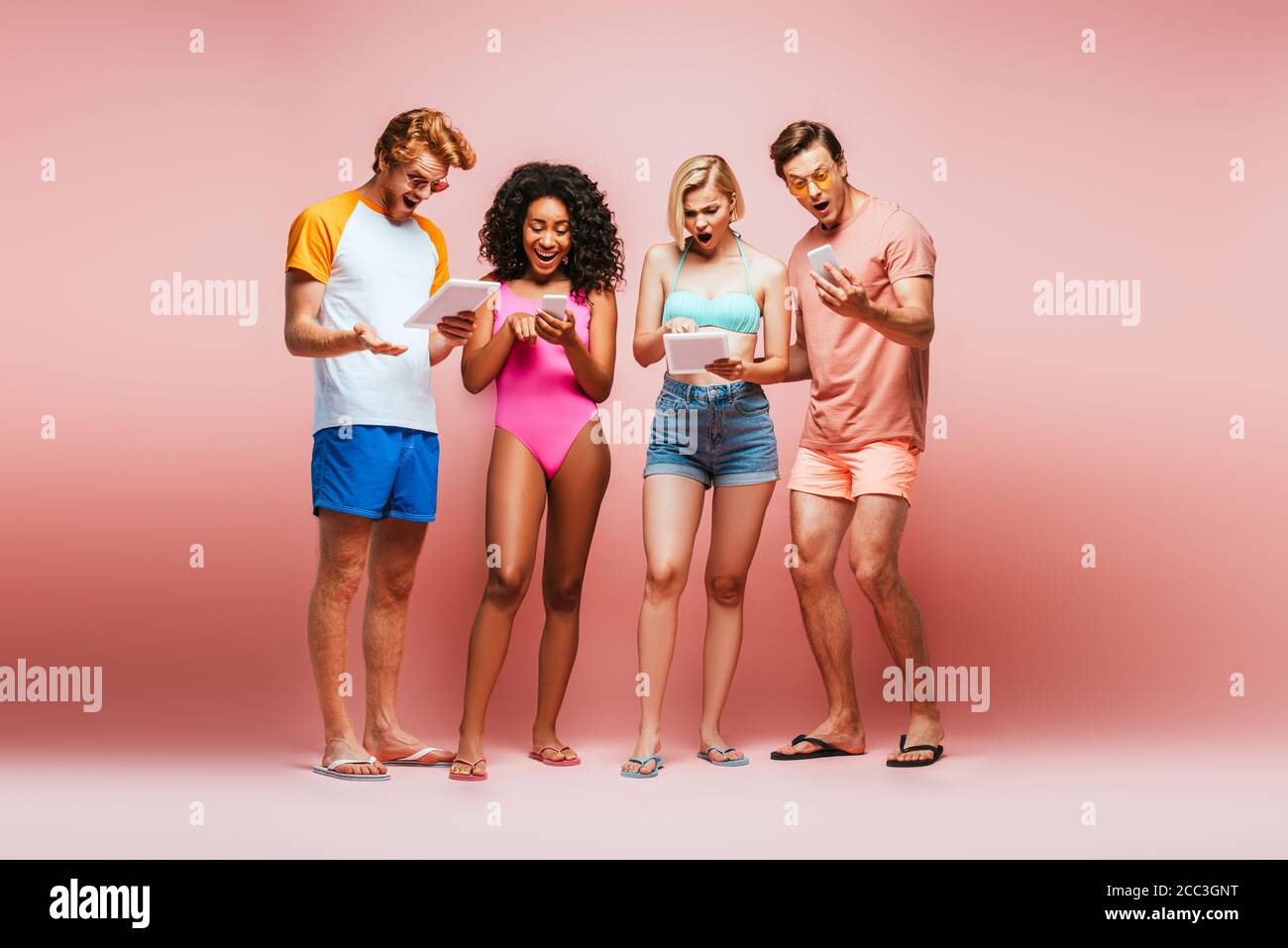 surprised multicultural friends in summer outfit using digital devices on pink Stock Photo