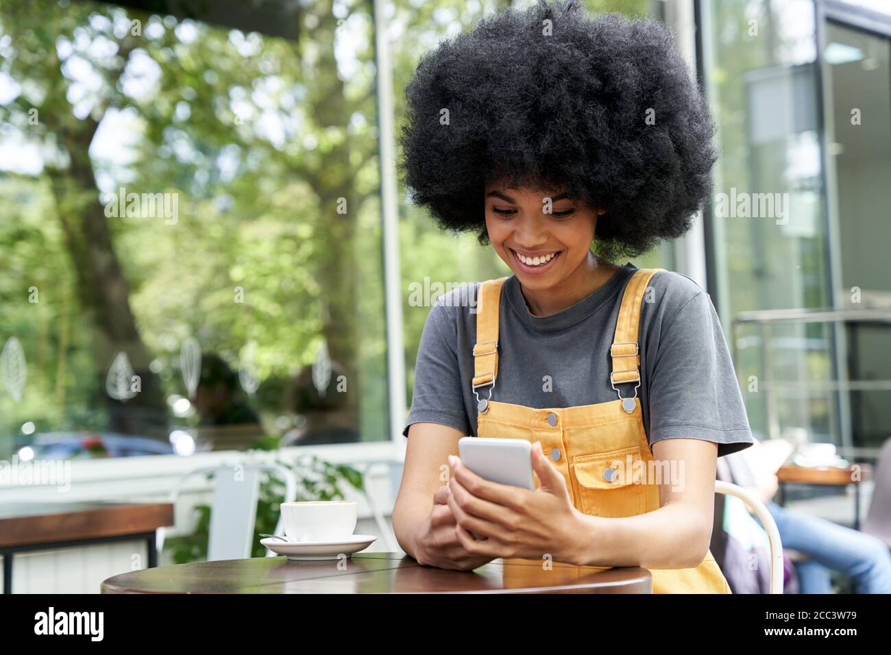 Smiling African hipster teen girl using phone sitting at outdoor cafe table. Stock Photo