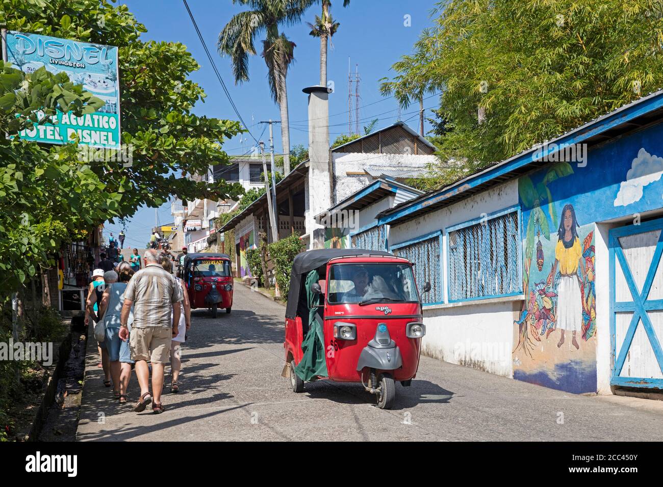 Western elderly tourists and red coloured tuk-tuks / mototaxis in street in the town Livingston, Izabal Department, Guatemala, Central America Stock Photo