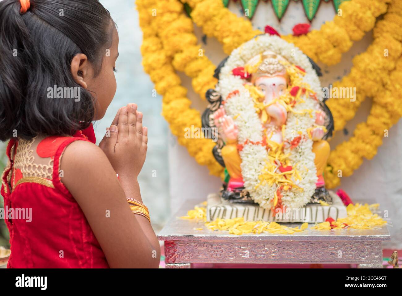 Girl Kid praying by closing eyes and folding hands in front of Lord Ganesha Idol during Ganesha or vinayaka Chaturthi festival ceremony at home - Stock Photo