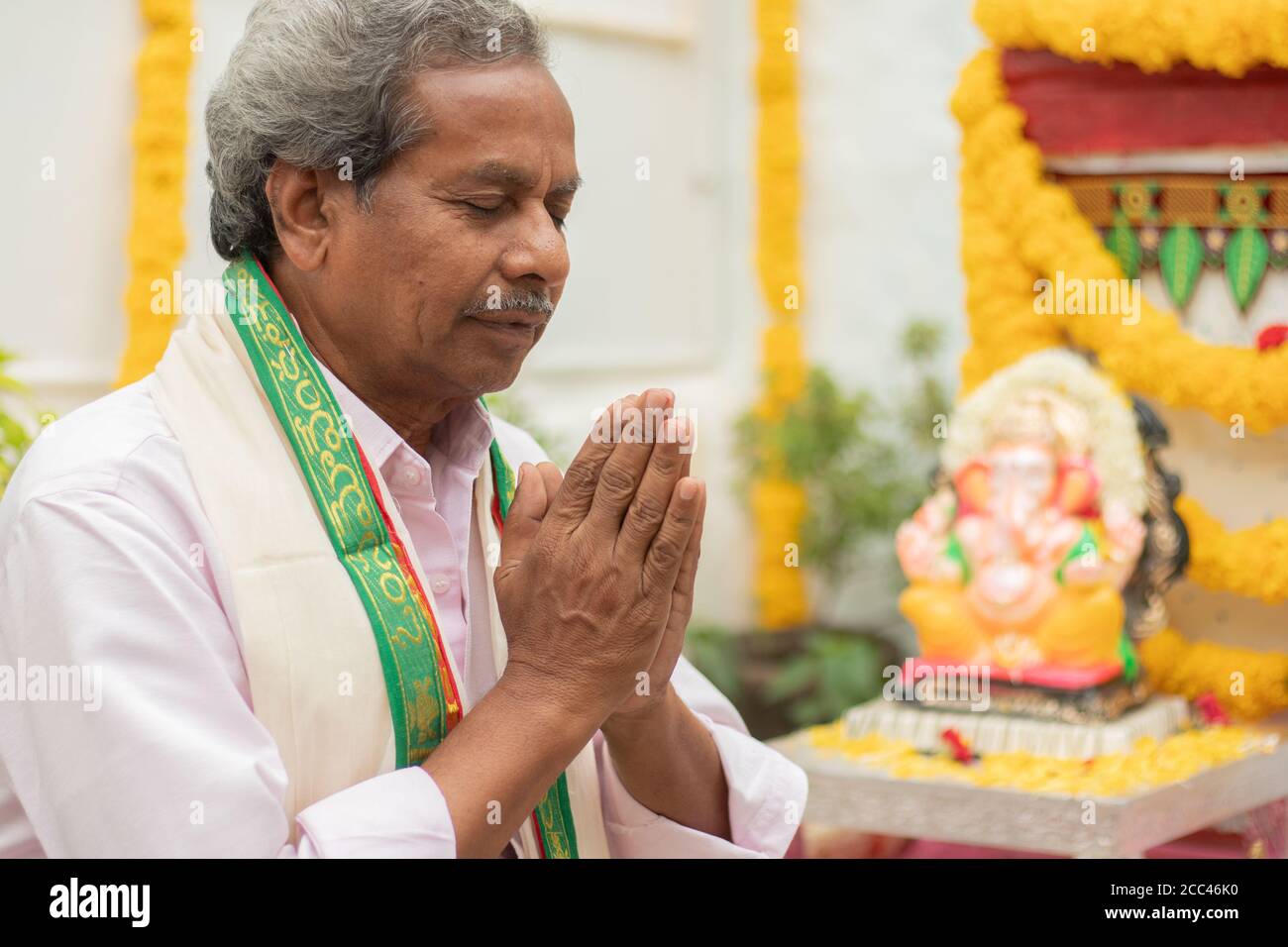 Elder man offering Prayer by namaste gesture in front of Lord Ganesha Idol during Ganesha or vinayaka Chaturthi festival ceremony at home. Stock Photo