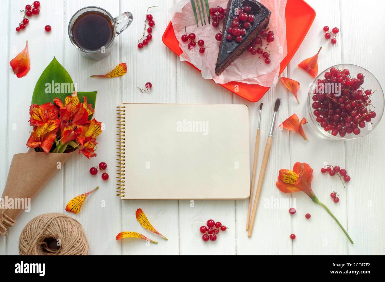 Open empty notebook with cup of coffee, red current cake slice, brushes and bouquet of flowers on the white wooden background, copy space. Top view. Stock Photo