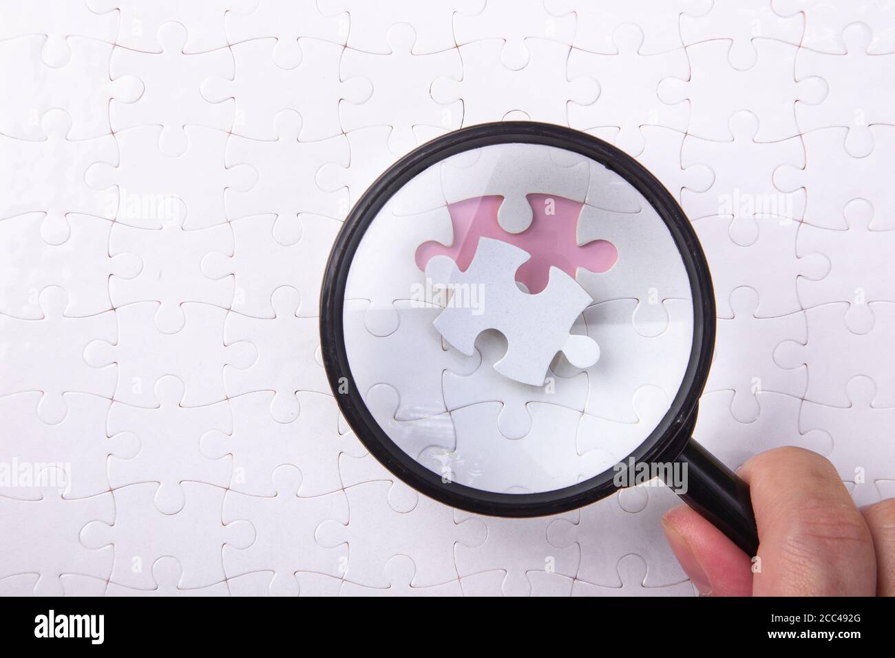 Hand holding a magnifying glass over a white jigsaw puzzle Stock Photo