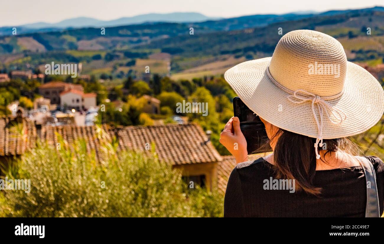 Gorgeous close-up view of a woman, wearing a big sun bonnet and taking photos with her mobile phone of the beautiful countryside of San Gimignano in... Stock Photo