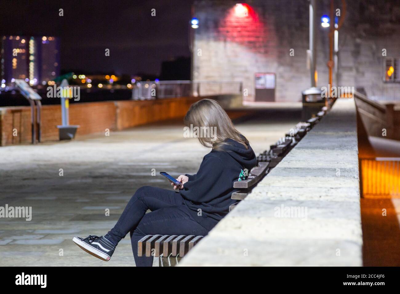 A woman sat alone on a bench at night looking at her mobile phone or cell phone Stock Photo